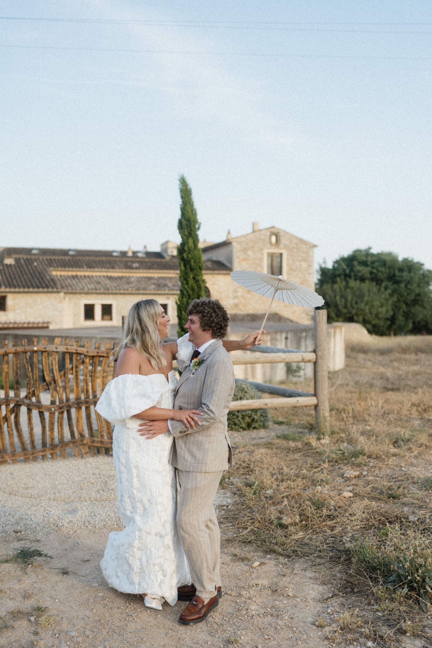 Bride and groom embrace outdoors at rustic Provence wedding venue, bride in off-shoulder gown, groom holding white parasol.