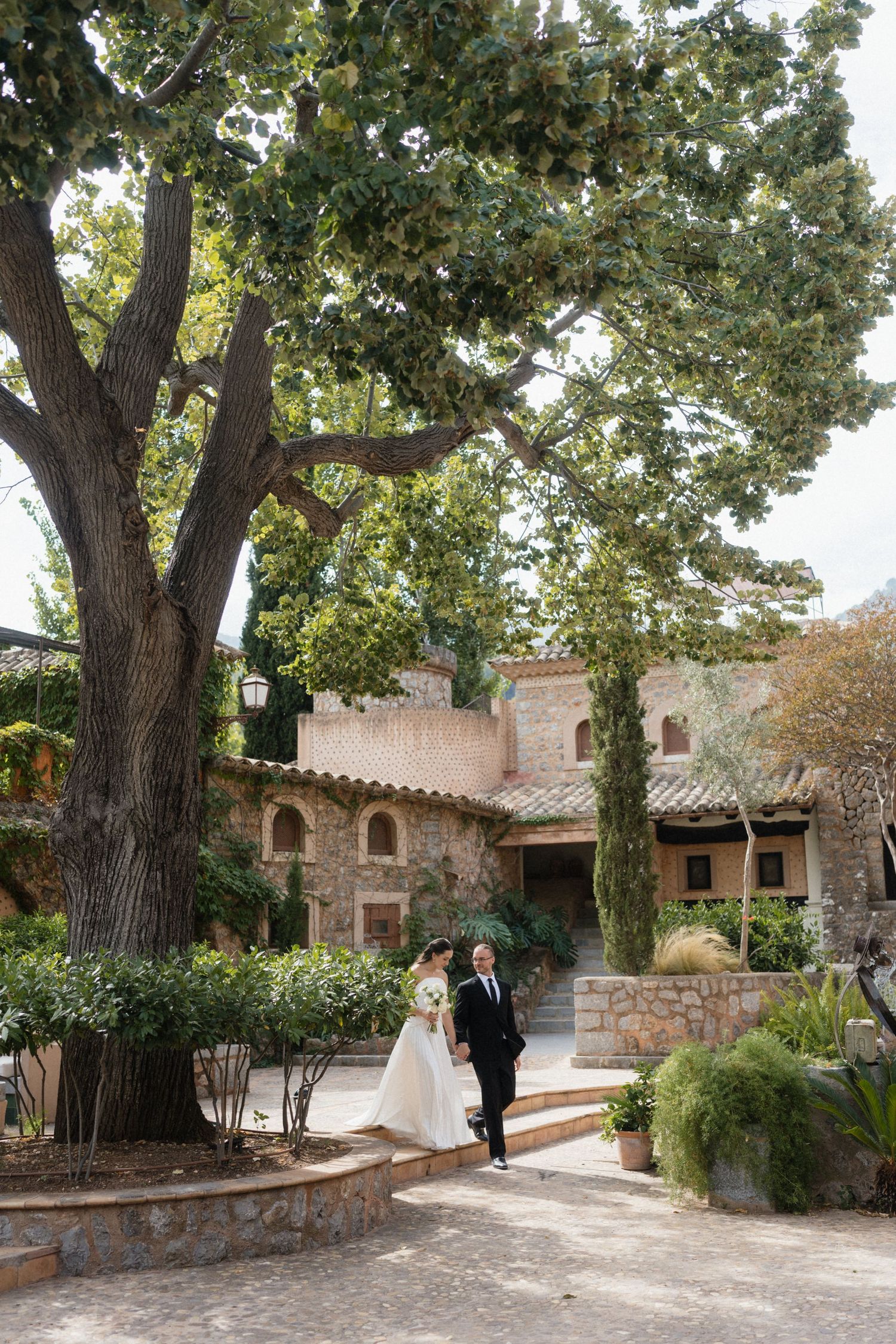 Bride in a white gown and groom in a black suit walk beneath a large old tree toward a rustic stone villa surrounded by lush gardens.