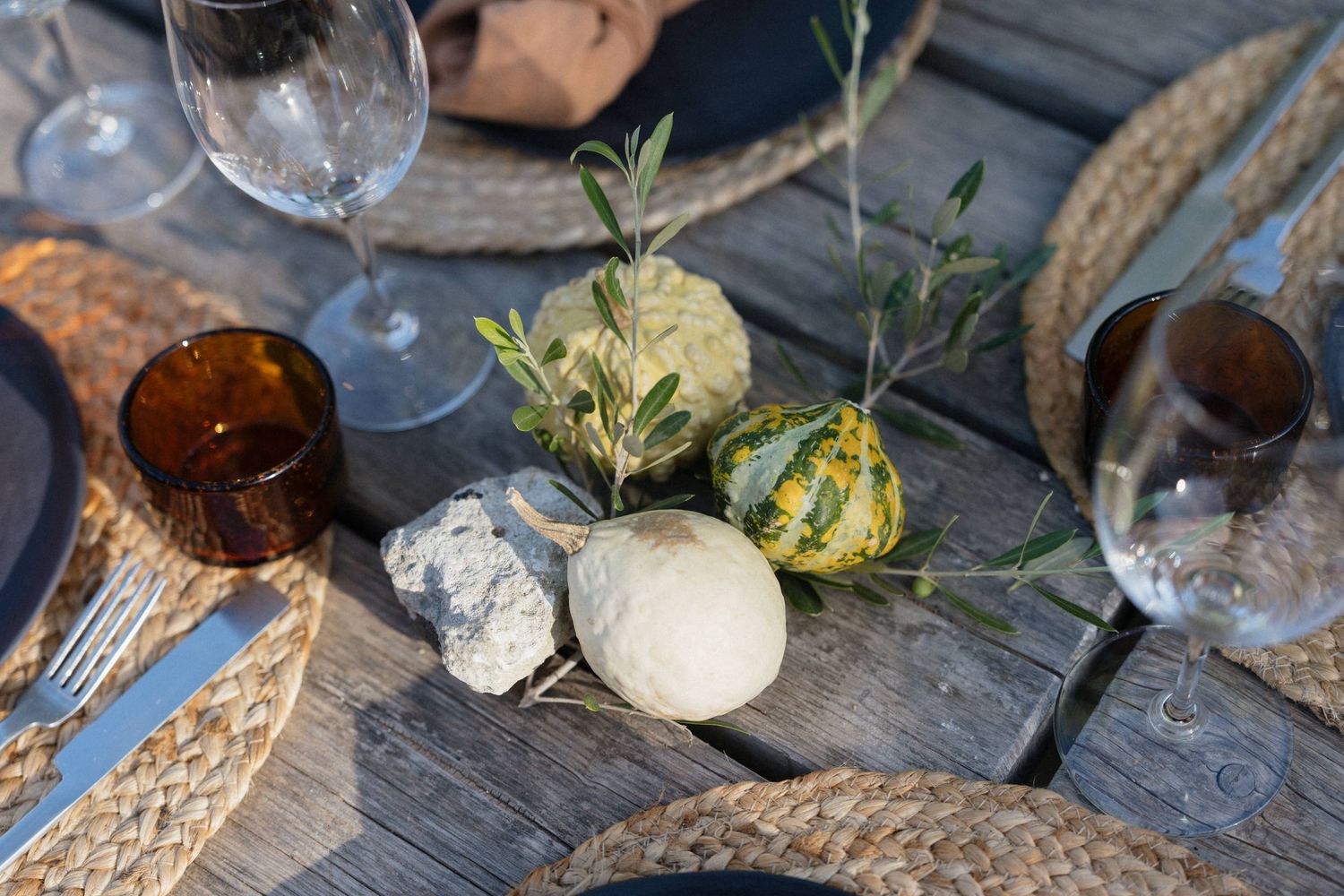 Rustic outdoor table setting with woven rattan placemats, wine glasses, amber tumbler, mini gourds, and olive sprigs on weathered wood.