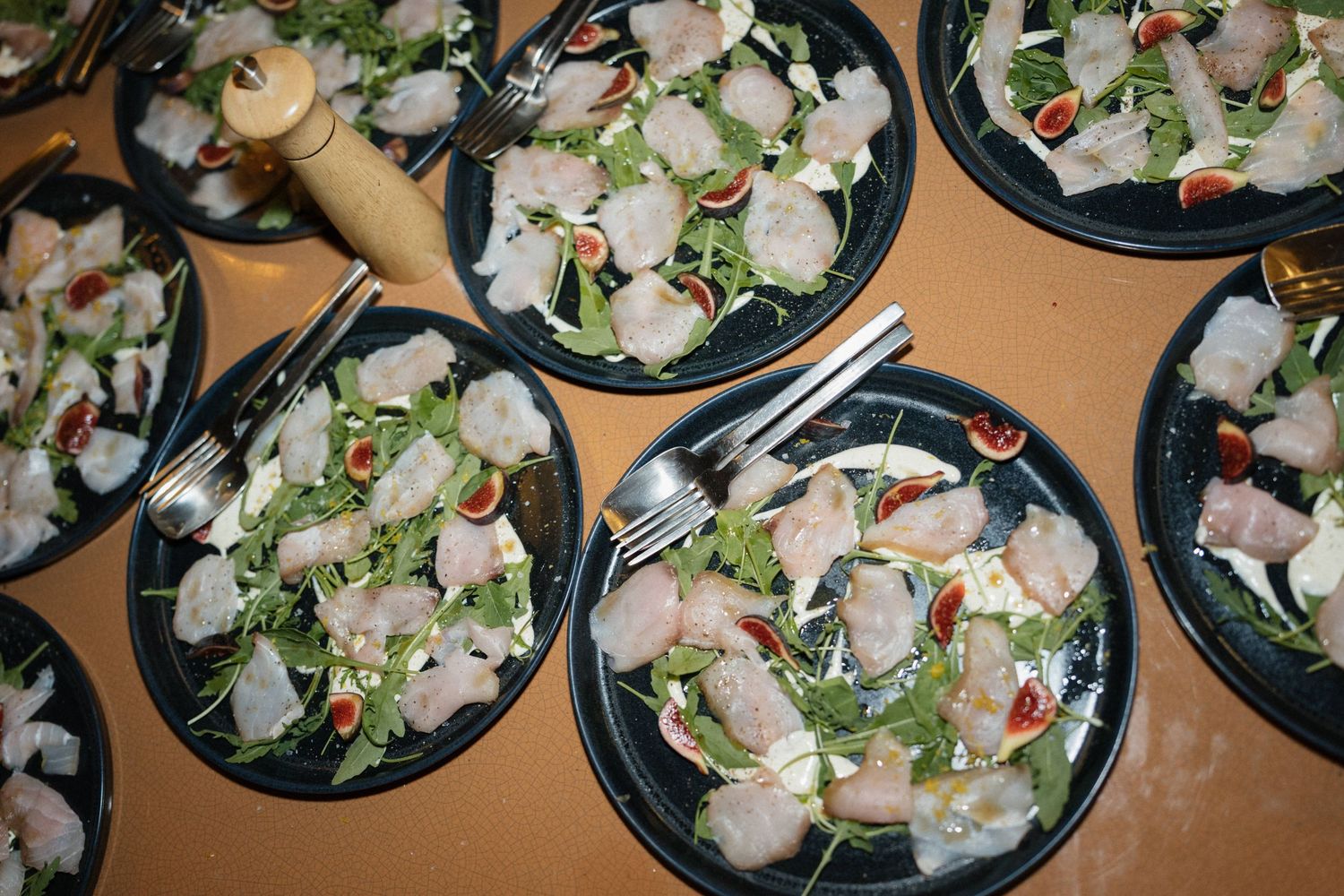 Black plates of fresh carpaccio topped with arugula and cherry tomatoes are arranged on a table with forks at a wedding dinner.