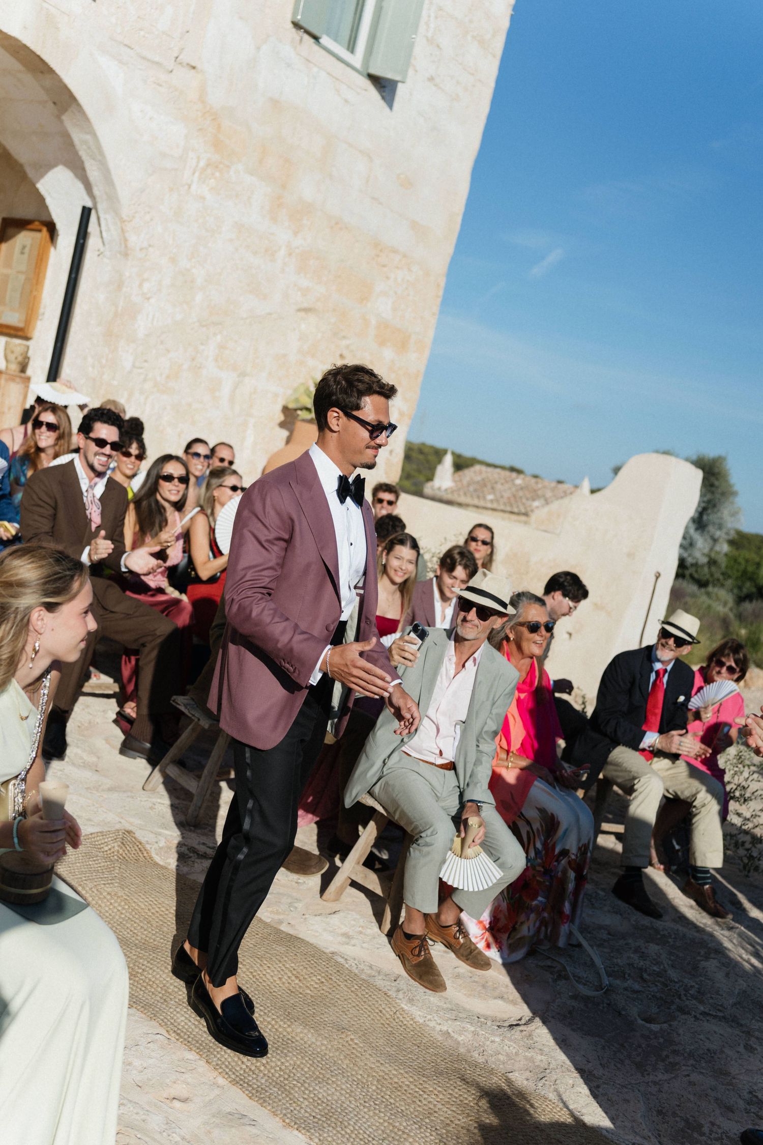 A groom in a burgundy blazer and bow tie walks down the aisle past smiling guests seated outdoors in the sunshine.