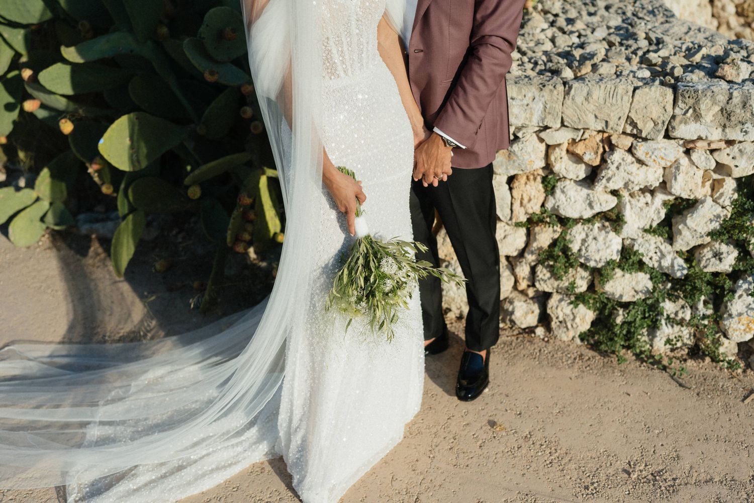 Bride and groom hold hands, with a green bouquet and long veil, beside a cactus and a stone wall.