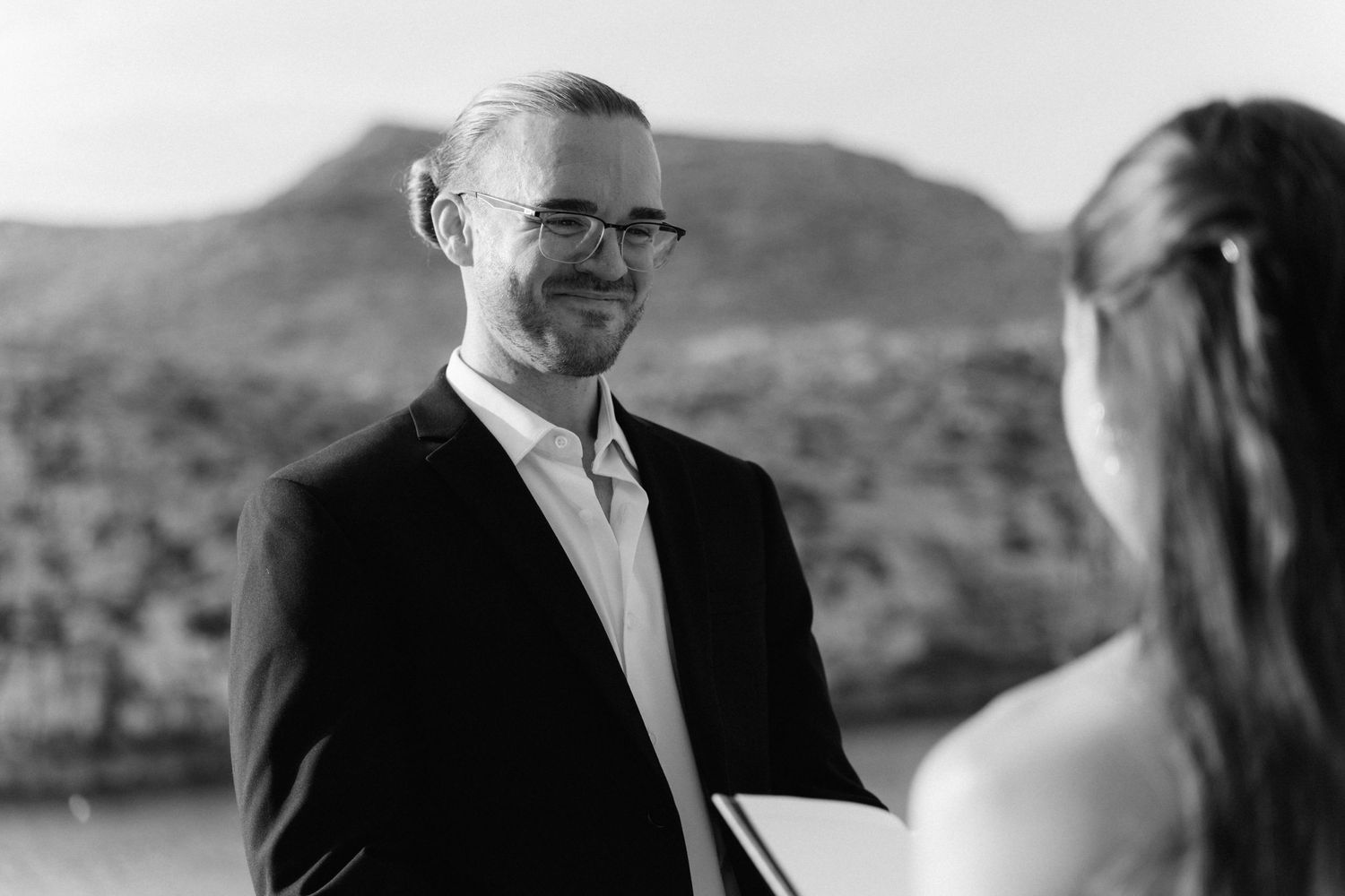 Smiling bearded groom in glasses and dark suit looks lovingly at his bride during an outdoor mountain wedding ceremony in black and white.