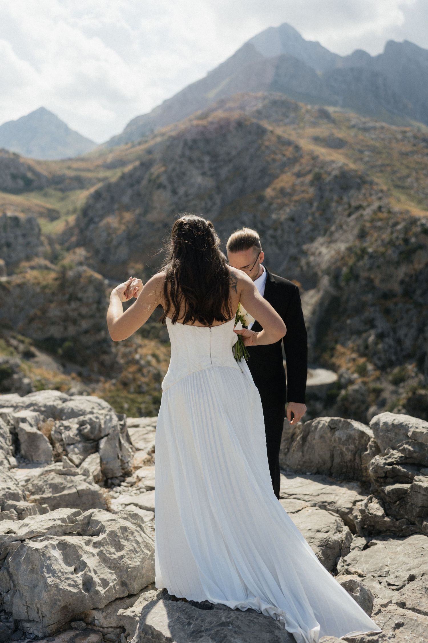 Bride in a flowing white gown and groom in a black suit embrace on rocky mountain terrain during an outdoor wedding photoshoot.