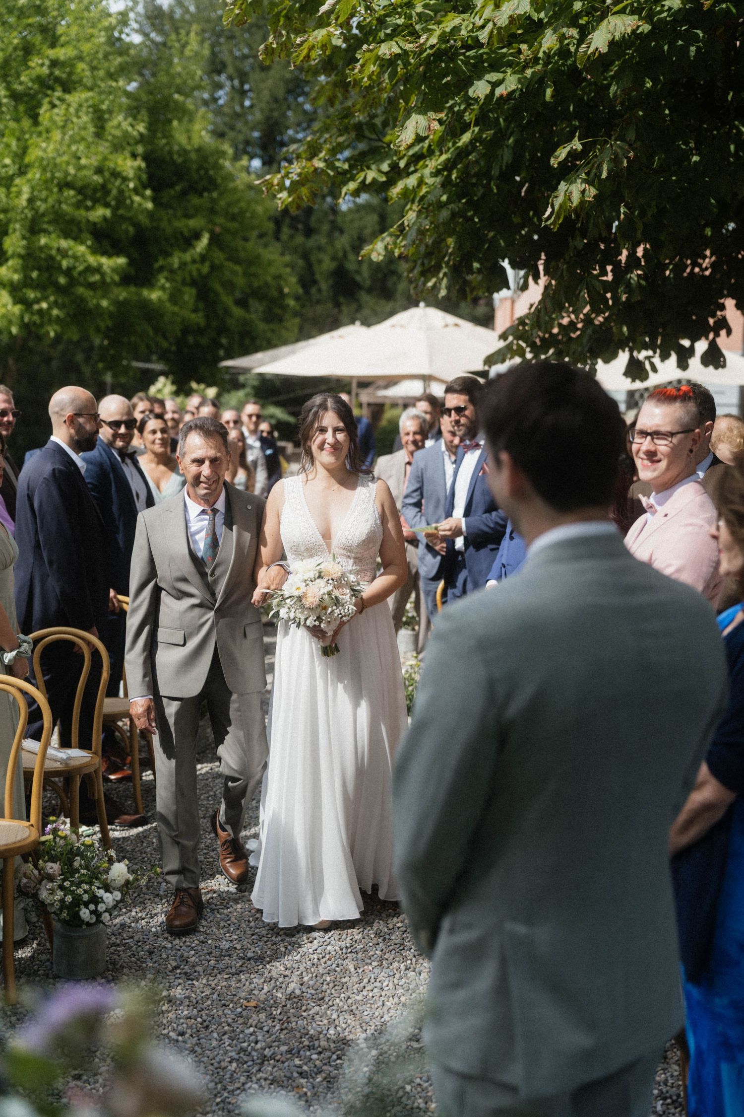 Bride walks down outdoor aisle in white gown holding bouquet, escorted by man in grey suit, guests lining the path.