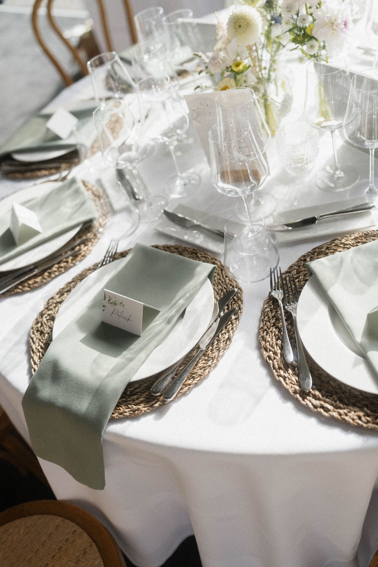 Wedding table setting with woven chargers, sage green napkins, crystal glasses, and white floral centerpieces.