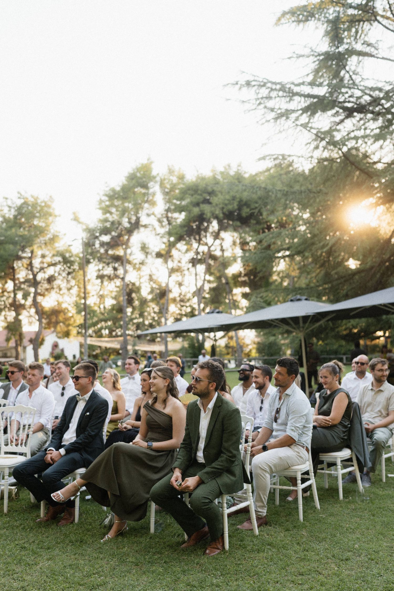 Wedding guests seated on white chairs outdoors at golden hour, surrounded by tall trees in a garden setting.