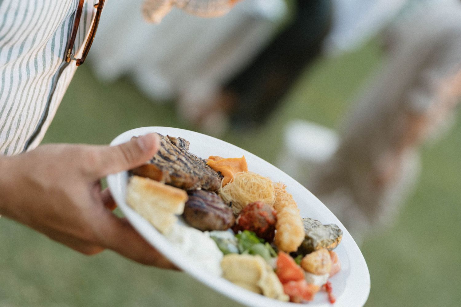 Person holding a white plate filled with grilled meat, fried foods, vegetables, and appetizers at an outdoor catered event.