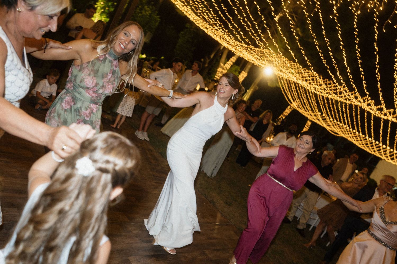 Bride in white gown dances joyfully with guests under twinkling fairy lights at an elegant outdoor wedding reception.