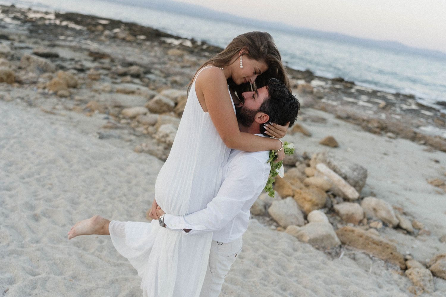 Couple in white embracing on a rocky beach, man lifting woman in a romantic seaside moment.