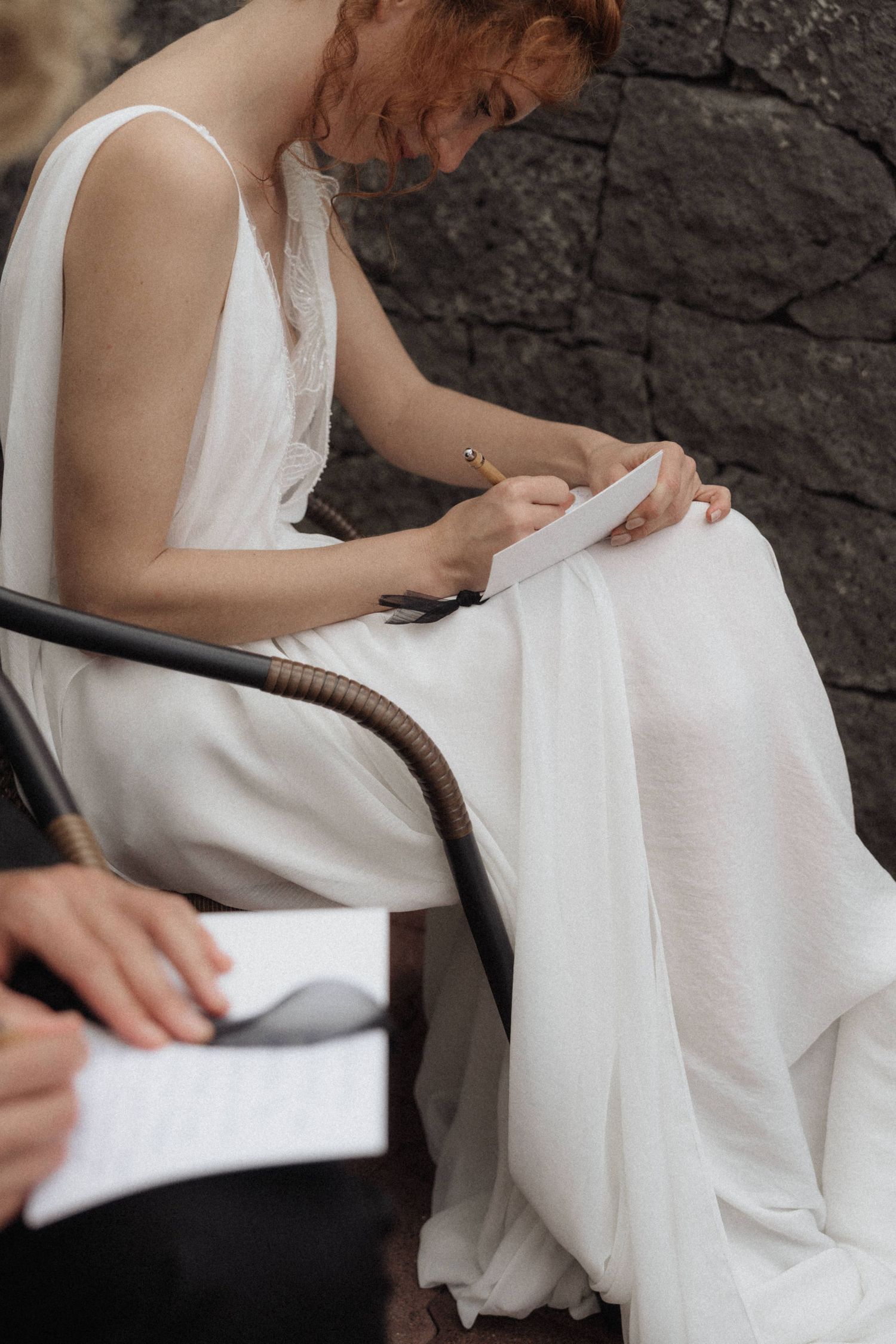 Bride in white flowing dress reads wedding vows while seated in rattan chair outdoors.
