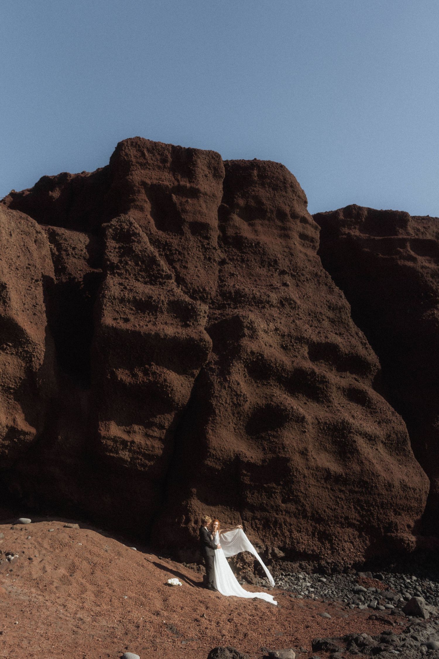 Bride in white gown stands before dramatic red volcanic rock cliffs on a black sand beach.