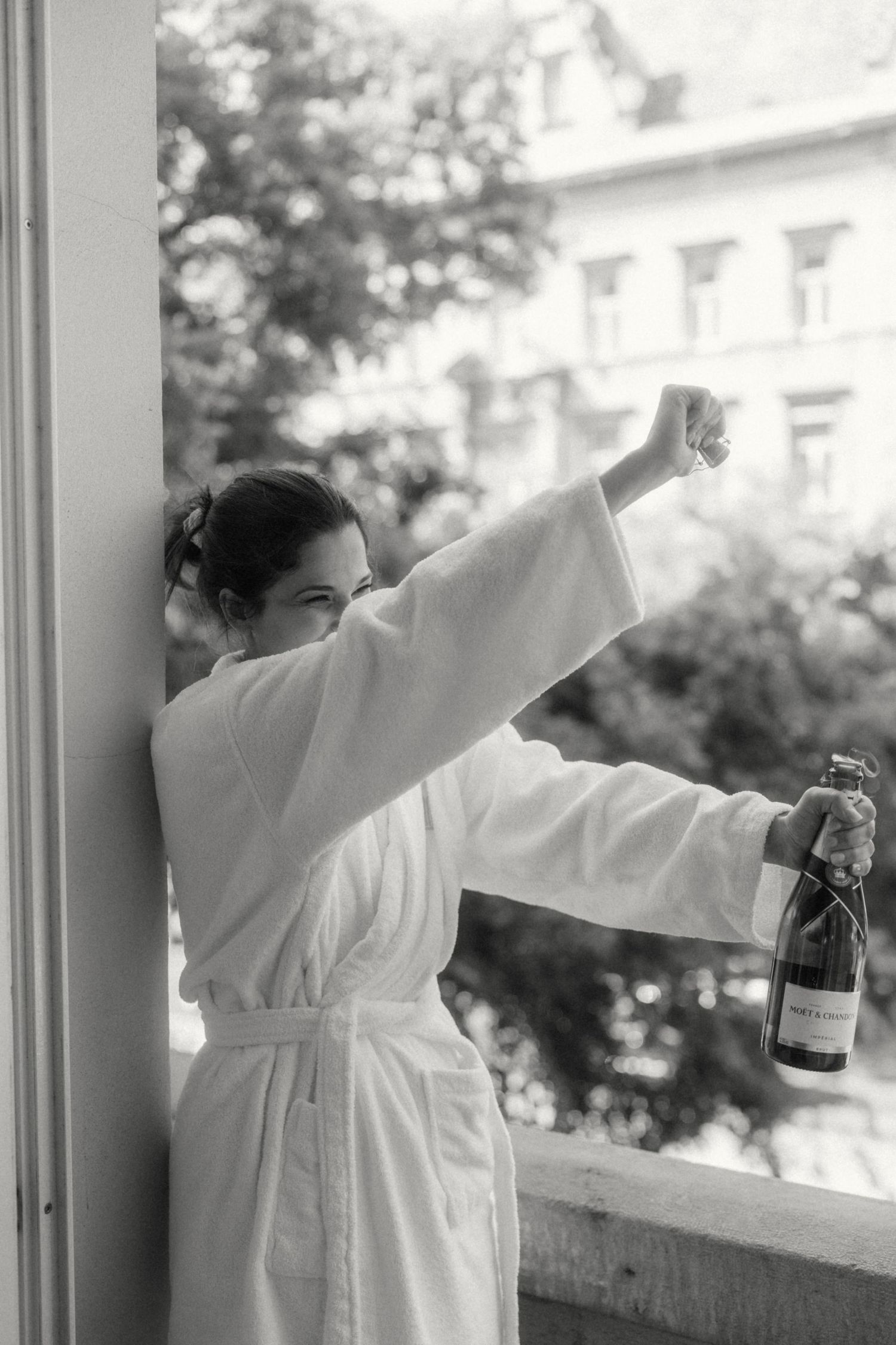 Woman in white bathrobe holding champagne bottle on balcony, black and white photo.