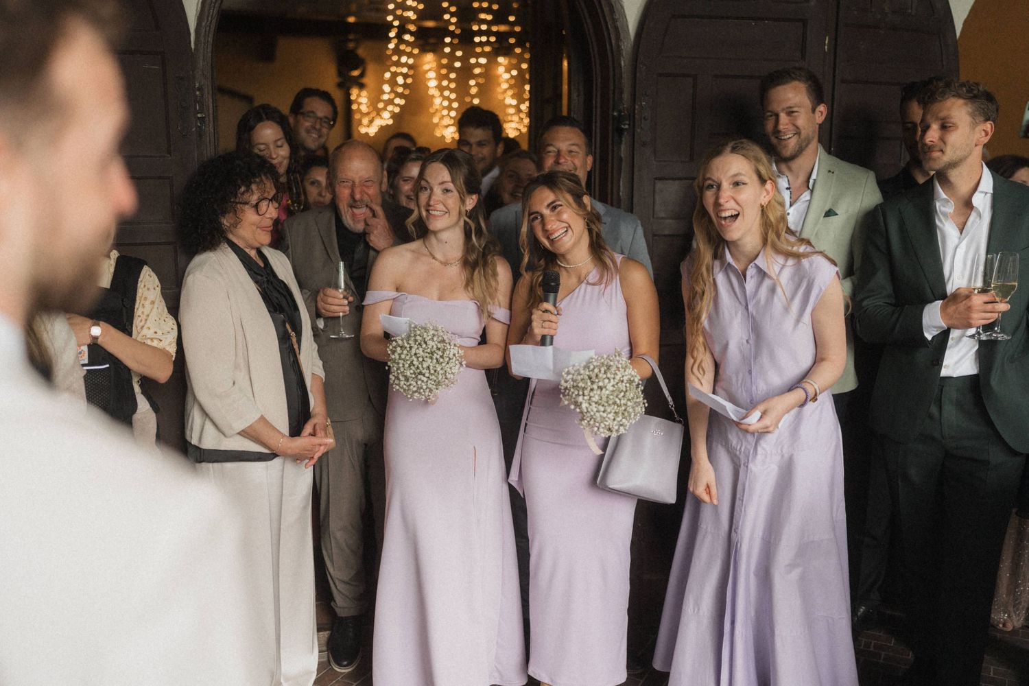 Three bridesmaids in matching lavender dresses holding floral bouquets reacting joyfully at a wedding.