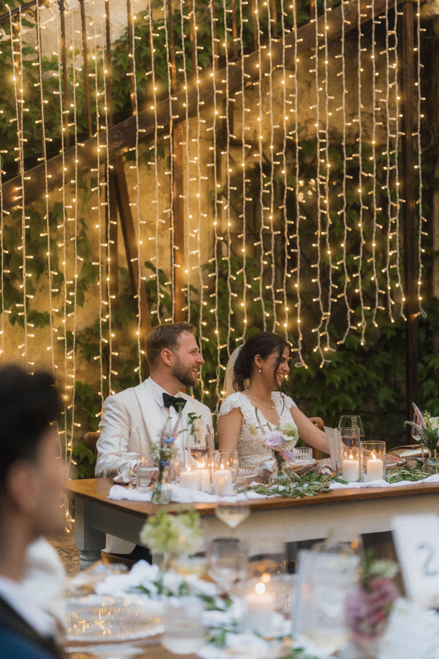Bride and groom smiling at a candlelit reception table with a glowing fairy light backdrop behind them.