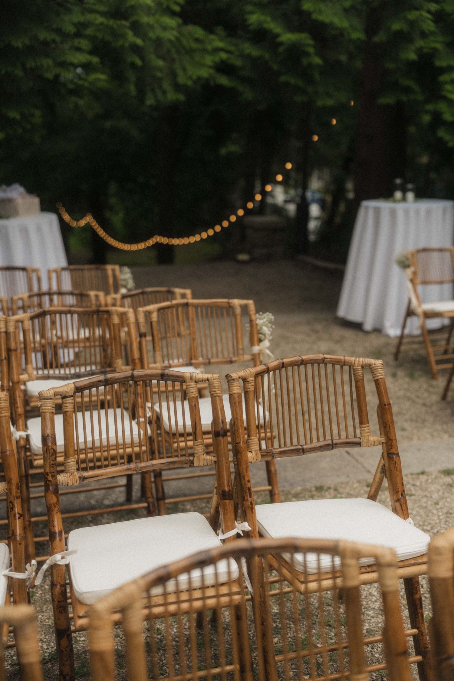 Rows of bamboo folding chairs with white cushions set up for an outdoor wedding ceremony with string lights.