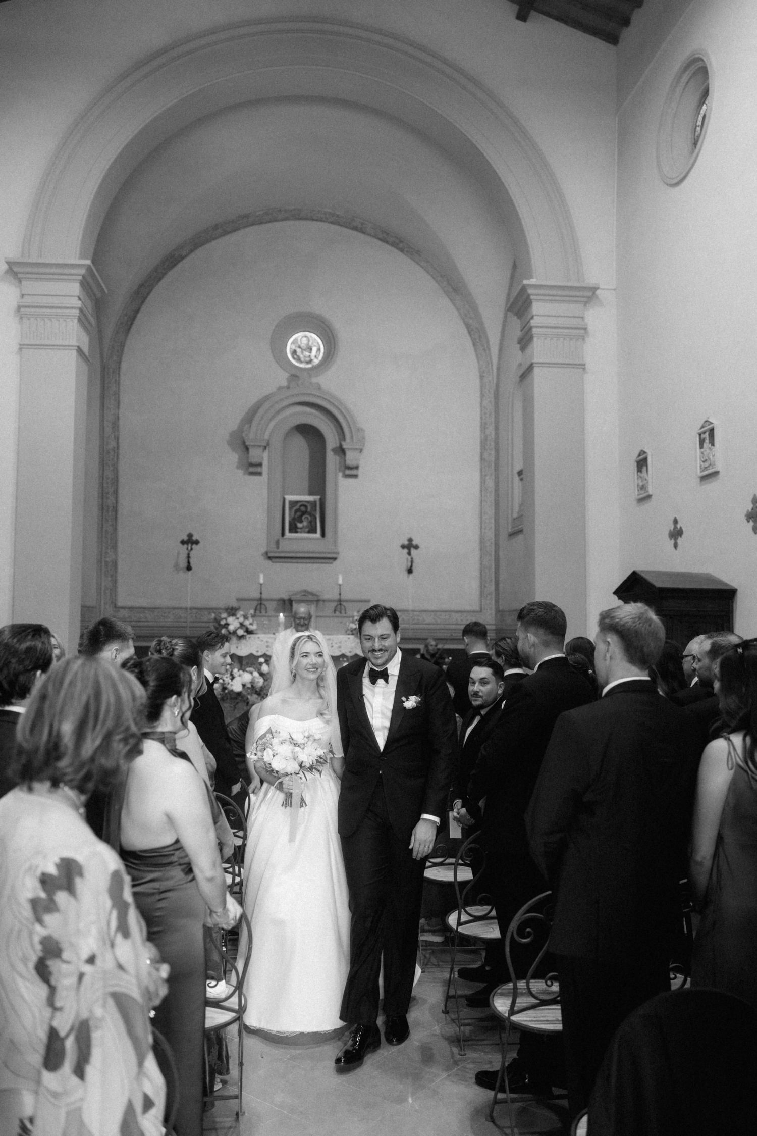 Bride and groom walk down church aisle after ceremony as guests celebrate in black and white photo.