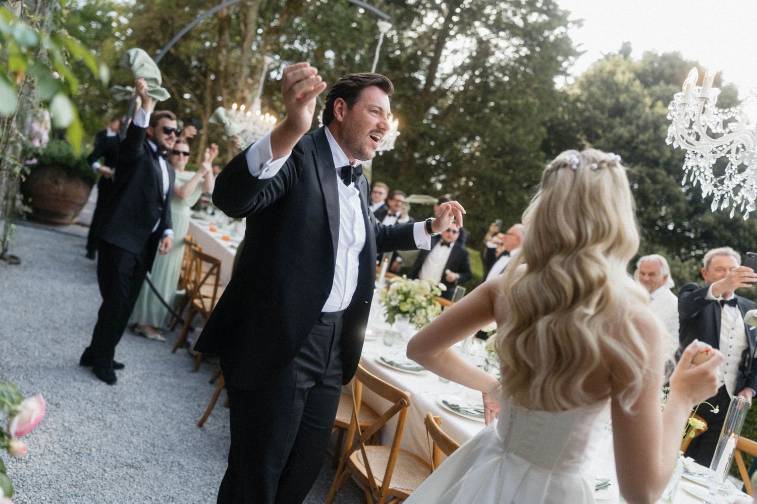 Joyful groom in tuxedo dancing and raising a glass with blonde bride at outdoor wedding reception.