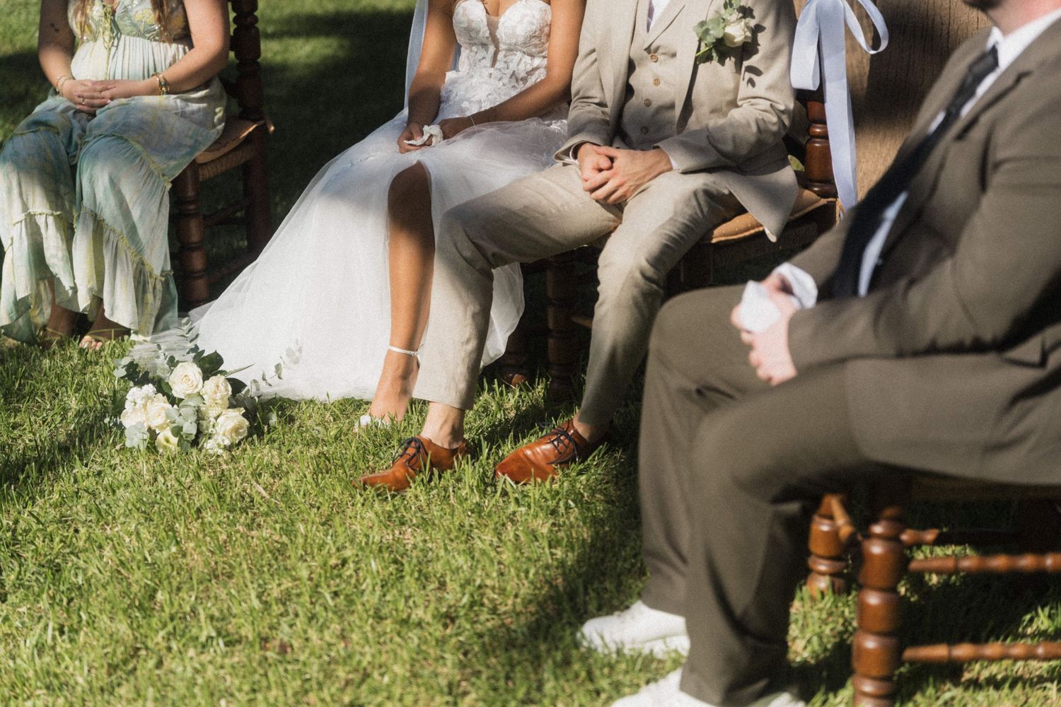 Close-up of bride in lace dress and groom in tan suit seated with wedding party on wooden chairs.
