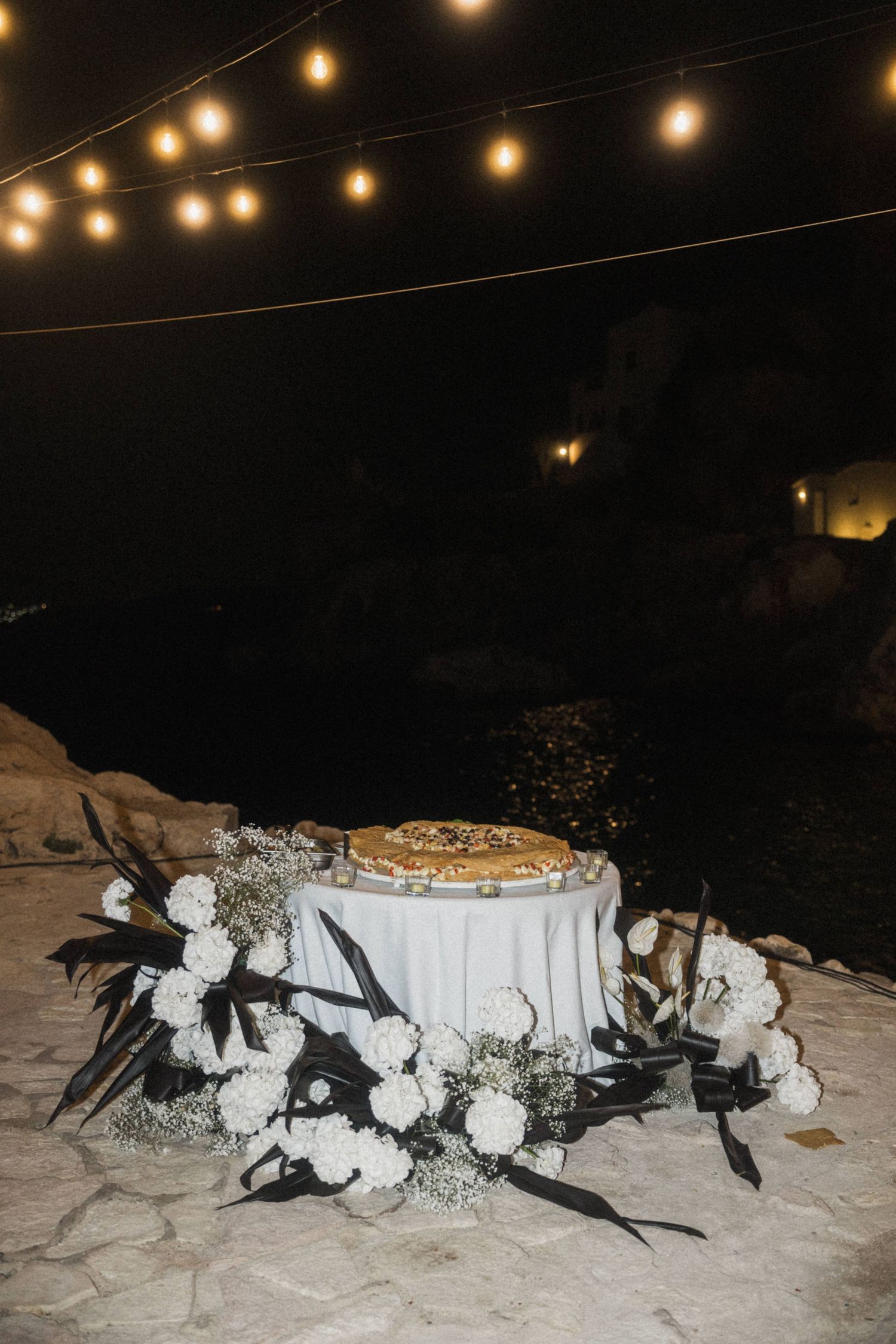 Elegant wedding pizza cake on white table with white floral decor under string lights at night.