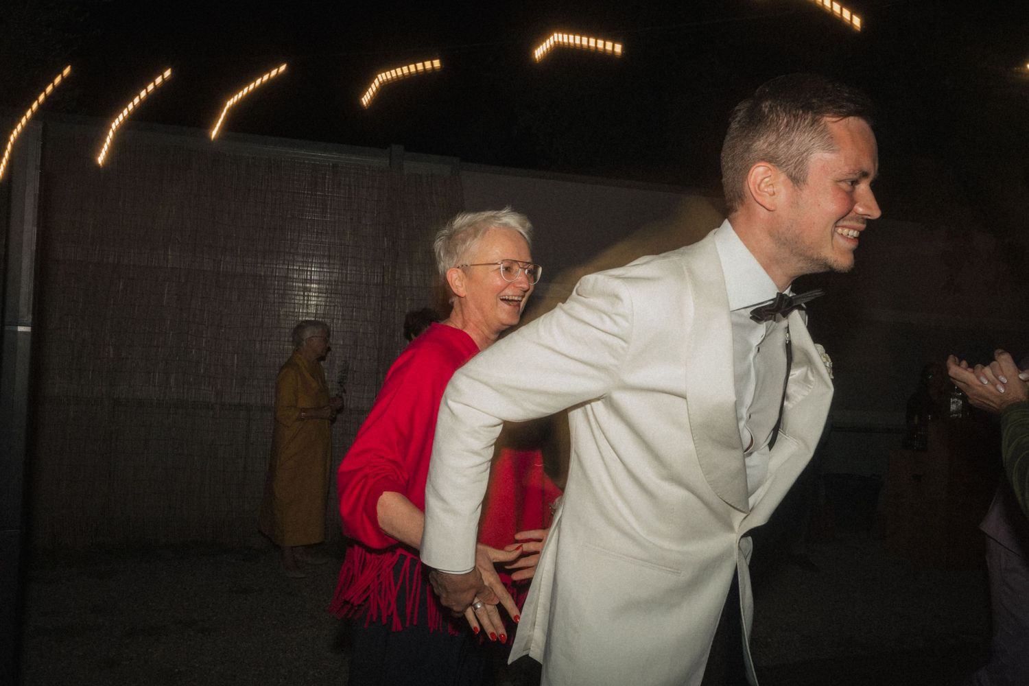 Groom in white tuxedo dances joyfully with older woman in red dress at outdoor evening reception.