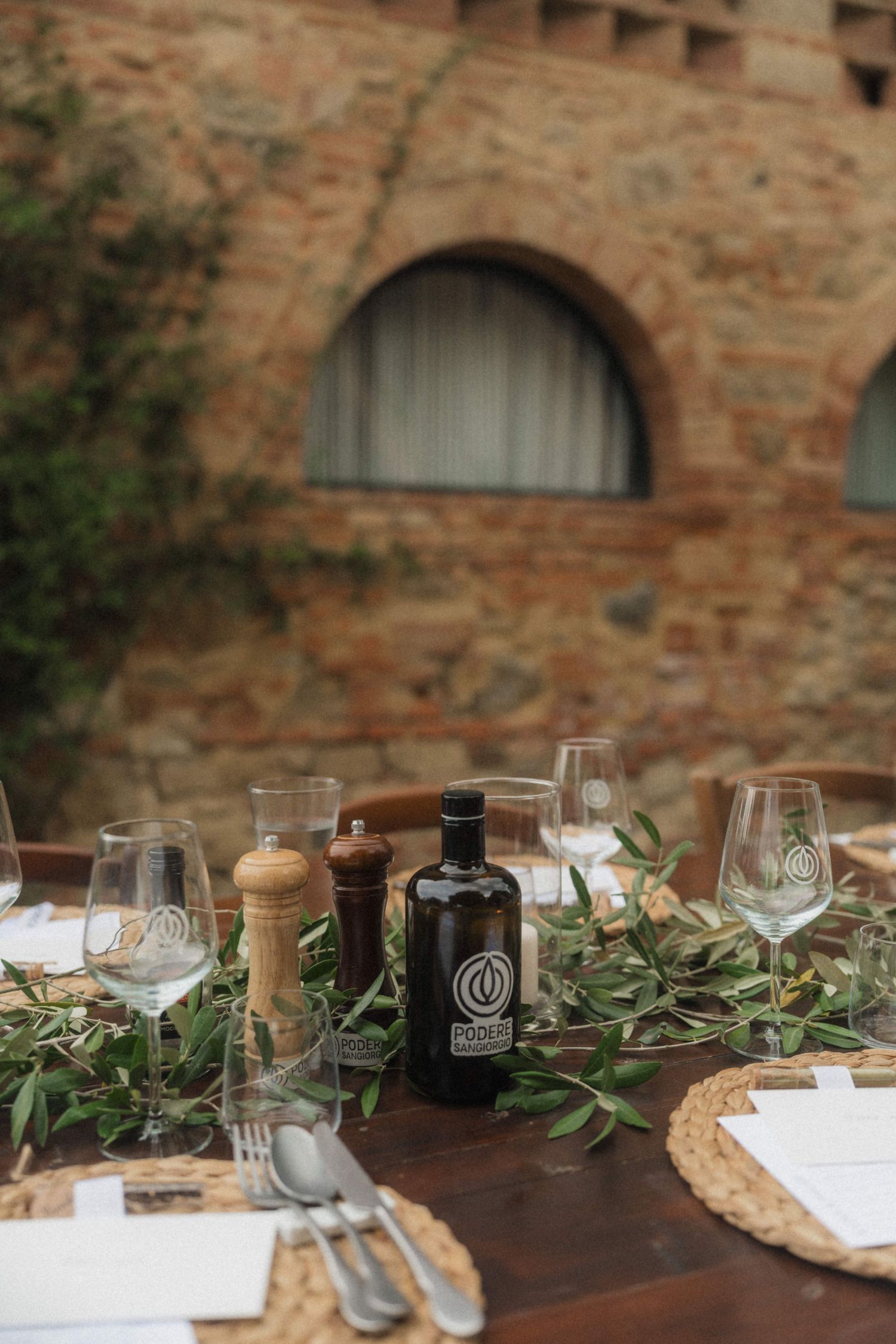 Elegant outdoor wedding table set with olive branches, wine glasses, and olive oil bottle at Tuscan venue.