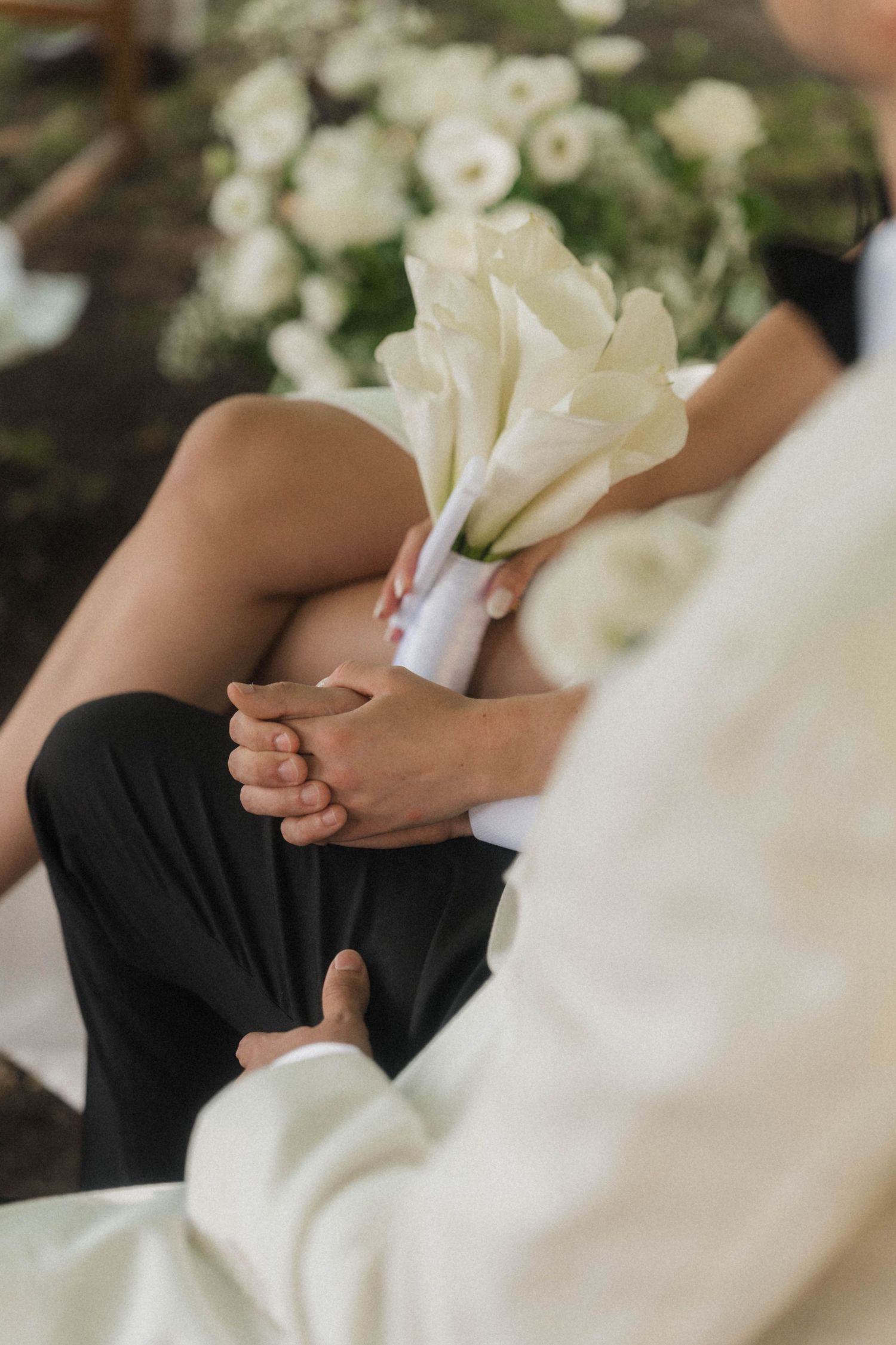 Bride holding white calla lily bouquet seated beside groom in black pants at elegant wedding ceremony.