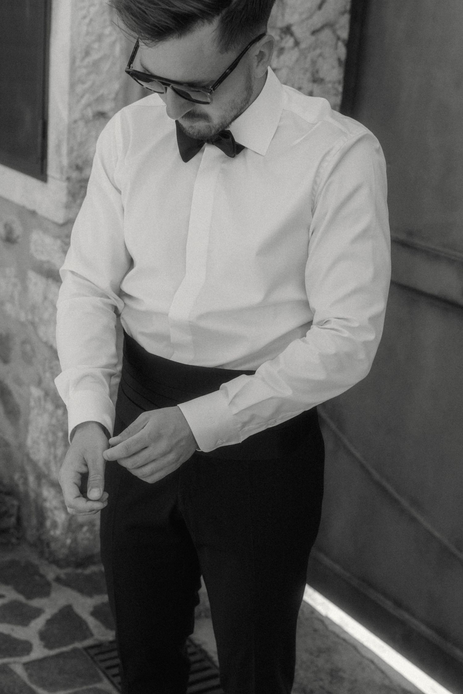 Elegant man in white tuxedo shirt and bow tie adjusting cufflinks in black and white photo.