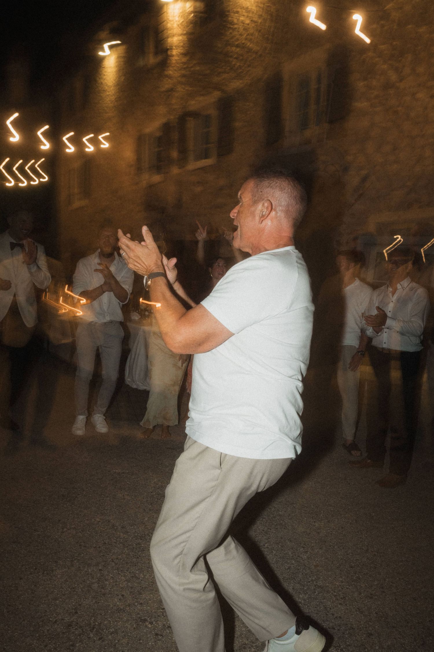 Man in white shirt dancing energetically at outdoor evening wedding reception with light trails.