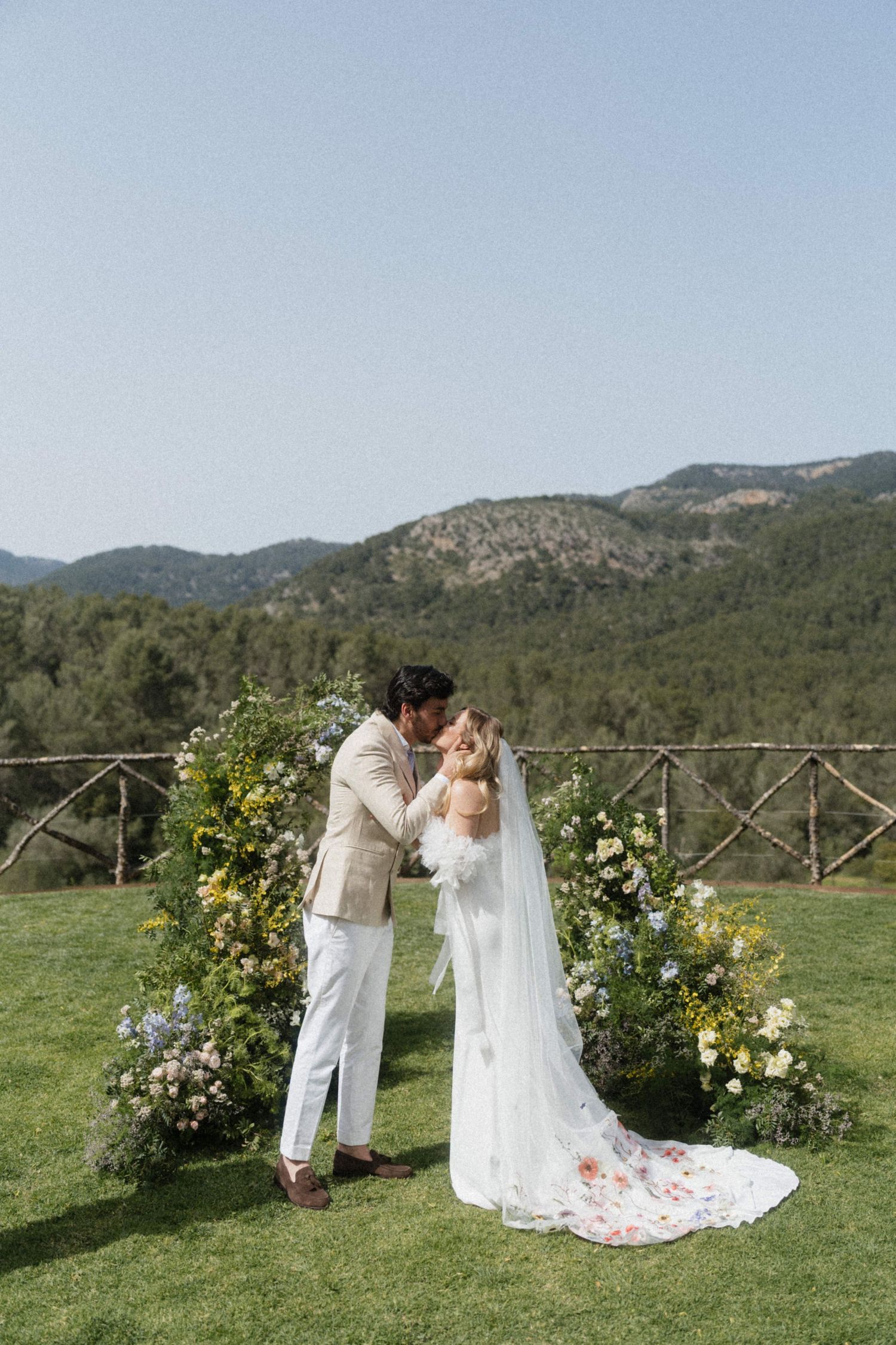 Bride and groom sharing first kiss at outdoor wedding altar surrounded by colorful wildflower arrangements.