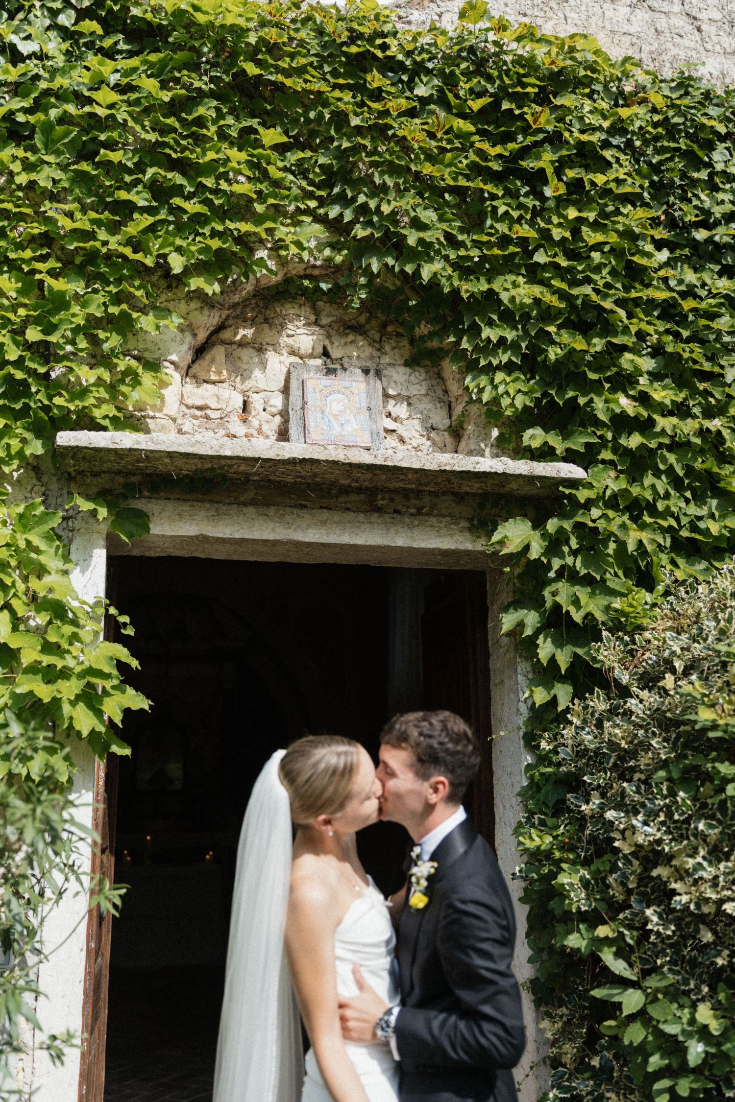 Newlywed couple shares a kiss in the ivy-covered stone doorway of a historic Italian chapel after their wedding.
