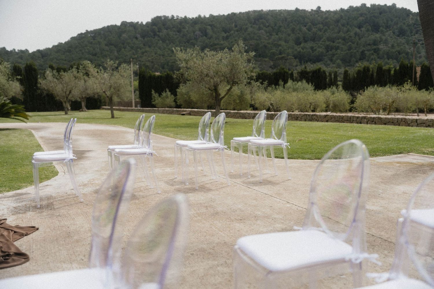 Elegant transparent ghost chairs arranged outdoors on a stone terrace with lush green lawn and olive trees in background.