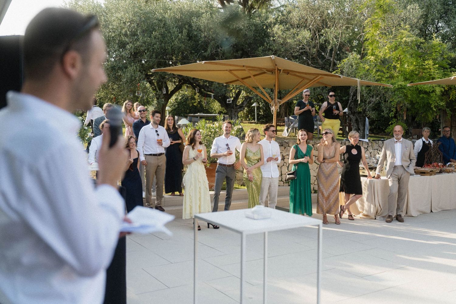 Outdoor wedding reception guests gathered on a patio under trees, listening to a speech near an elegant white table.