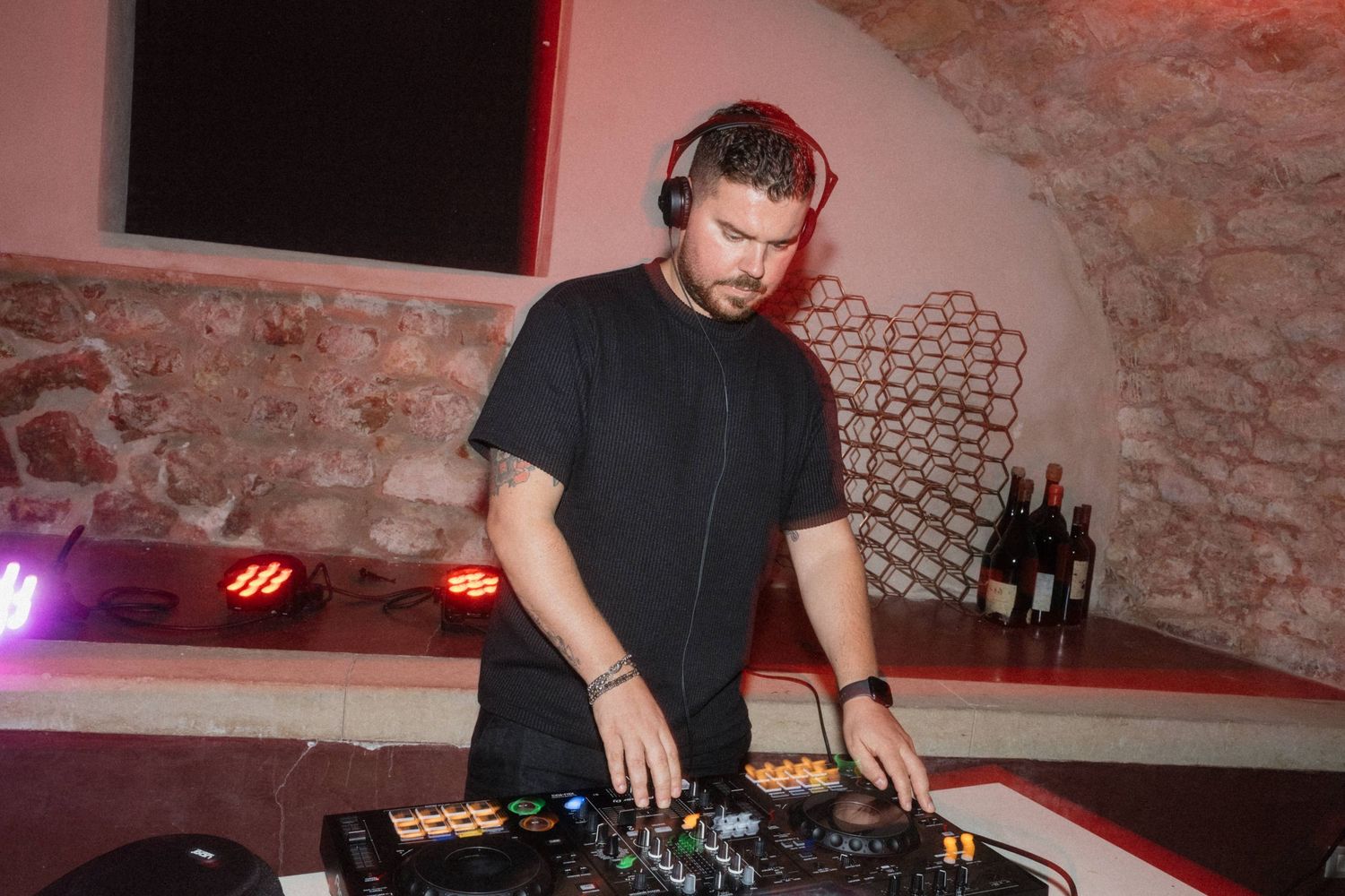 Male DJ wearing headphones mixing music on a DJ controller at an indoor event with red ambient lighting.
