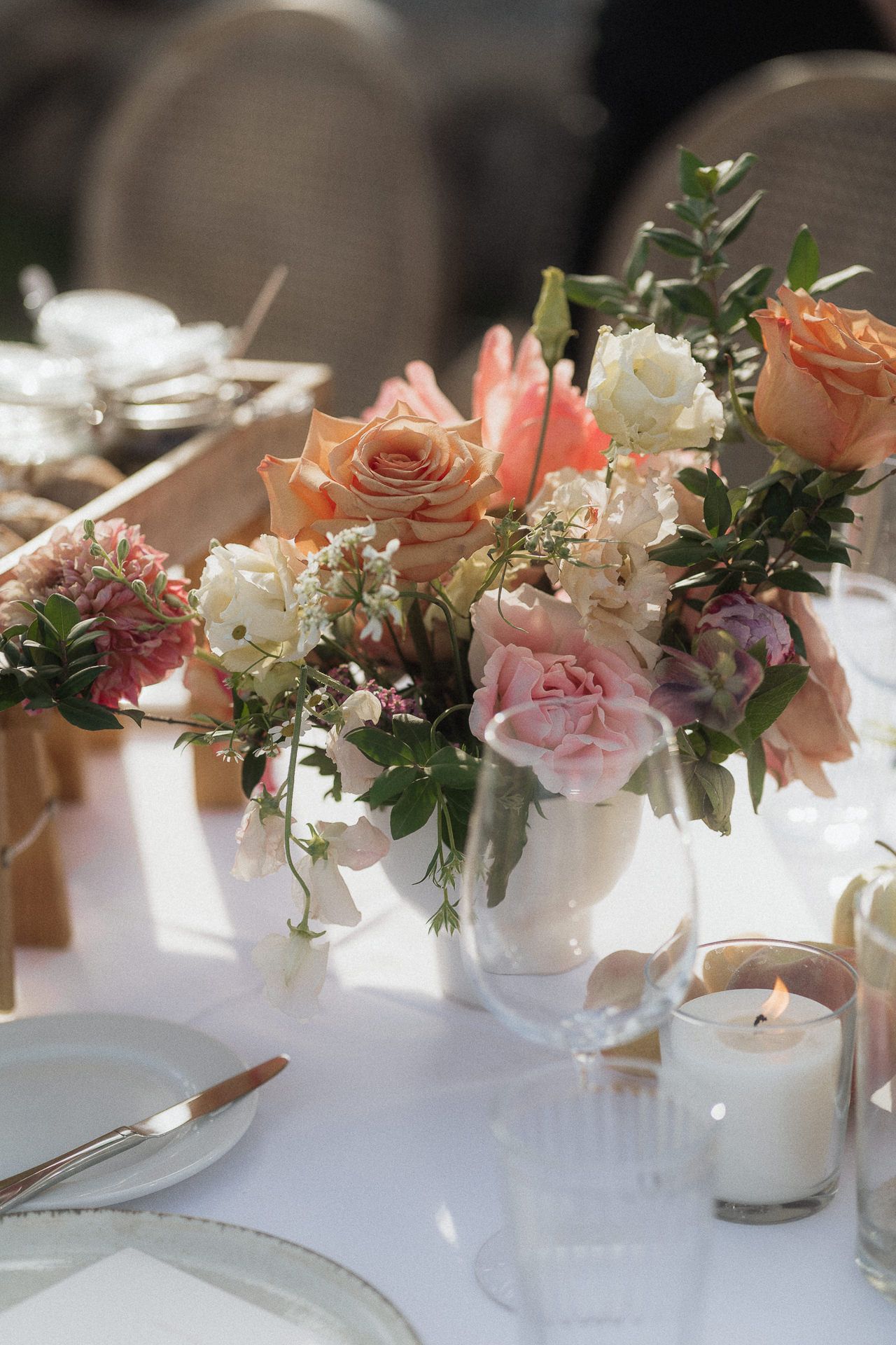 Elegant wedding table centerpiece with peach roses, pink peonies, and white flowers in a glass vase beside a lit candle.