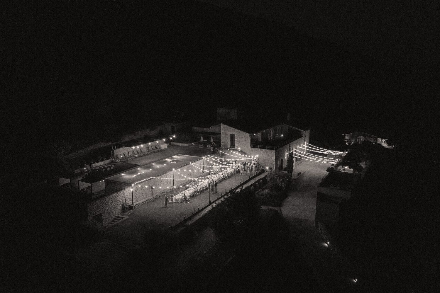 Aerial black and white night photo of an outdoor wedding venue glowing with string lights in a dark landscape.
