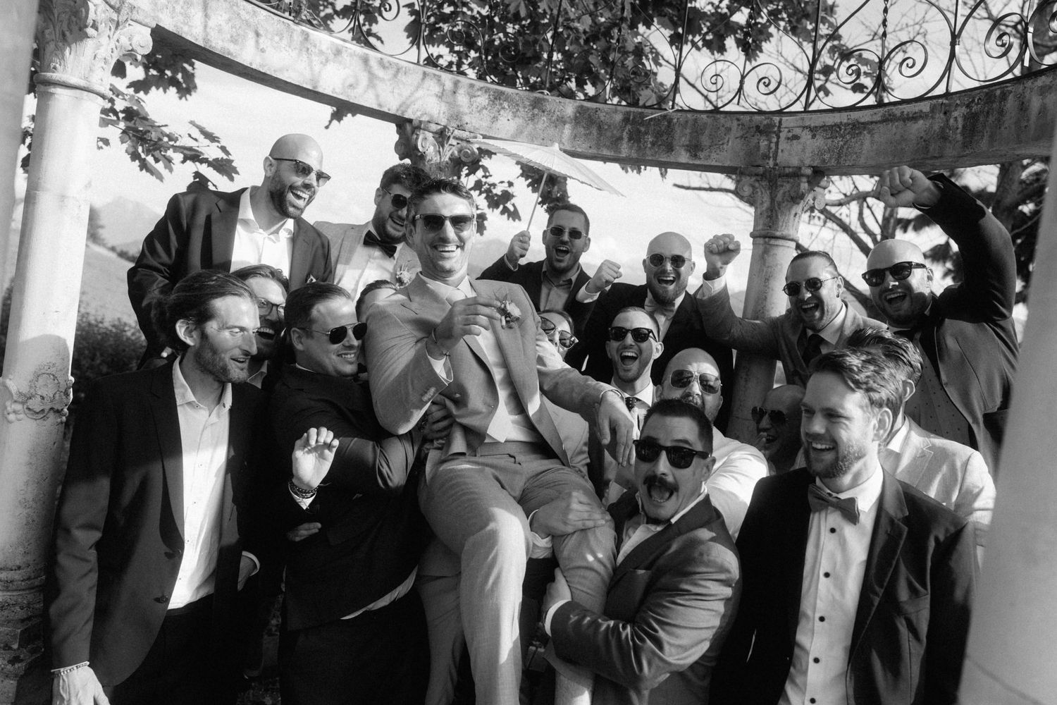 Black and white photo of groom lifted by cheering groomsmen near a decorative stone gazebo.