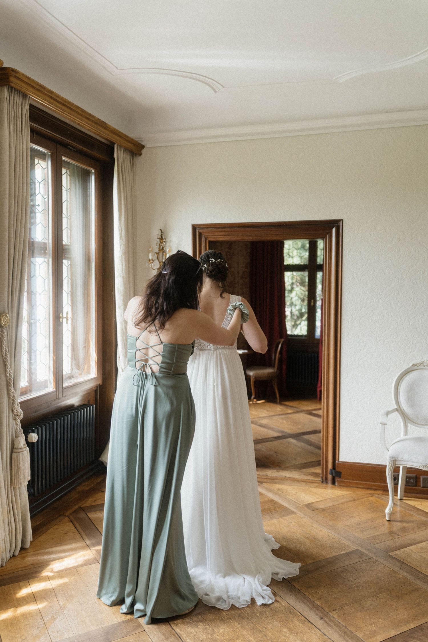 Bridesmaid in sage green gown helping bride in white wedding dress prepare in sunlit vintage room.