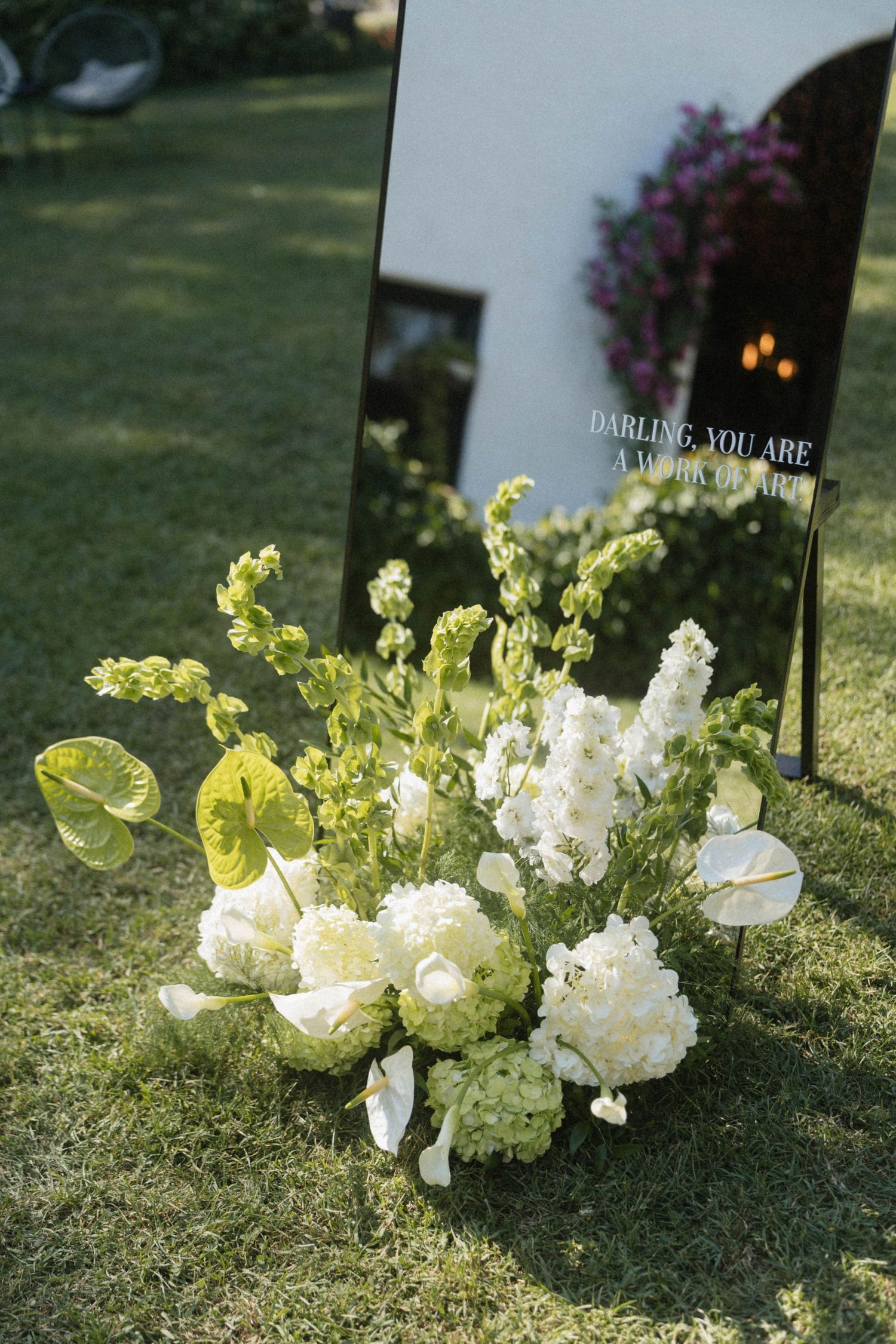 White floral arrangement with green foliage beside a mirror sign on grass at an outdoor wedding.