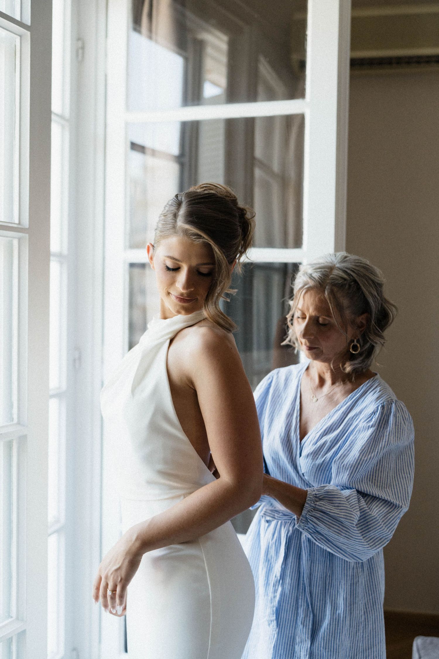 Mother helping bride zip up elegant white halter wedding dress by a bright window.