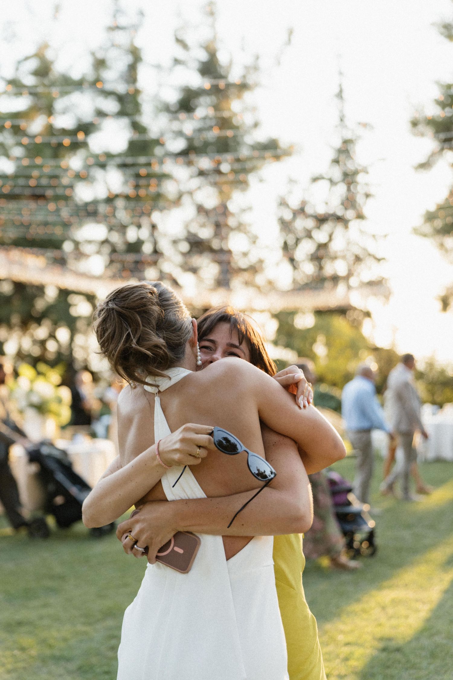 Two women share a warm embrace at an outdoor wedding reception on a sunny day.