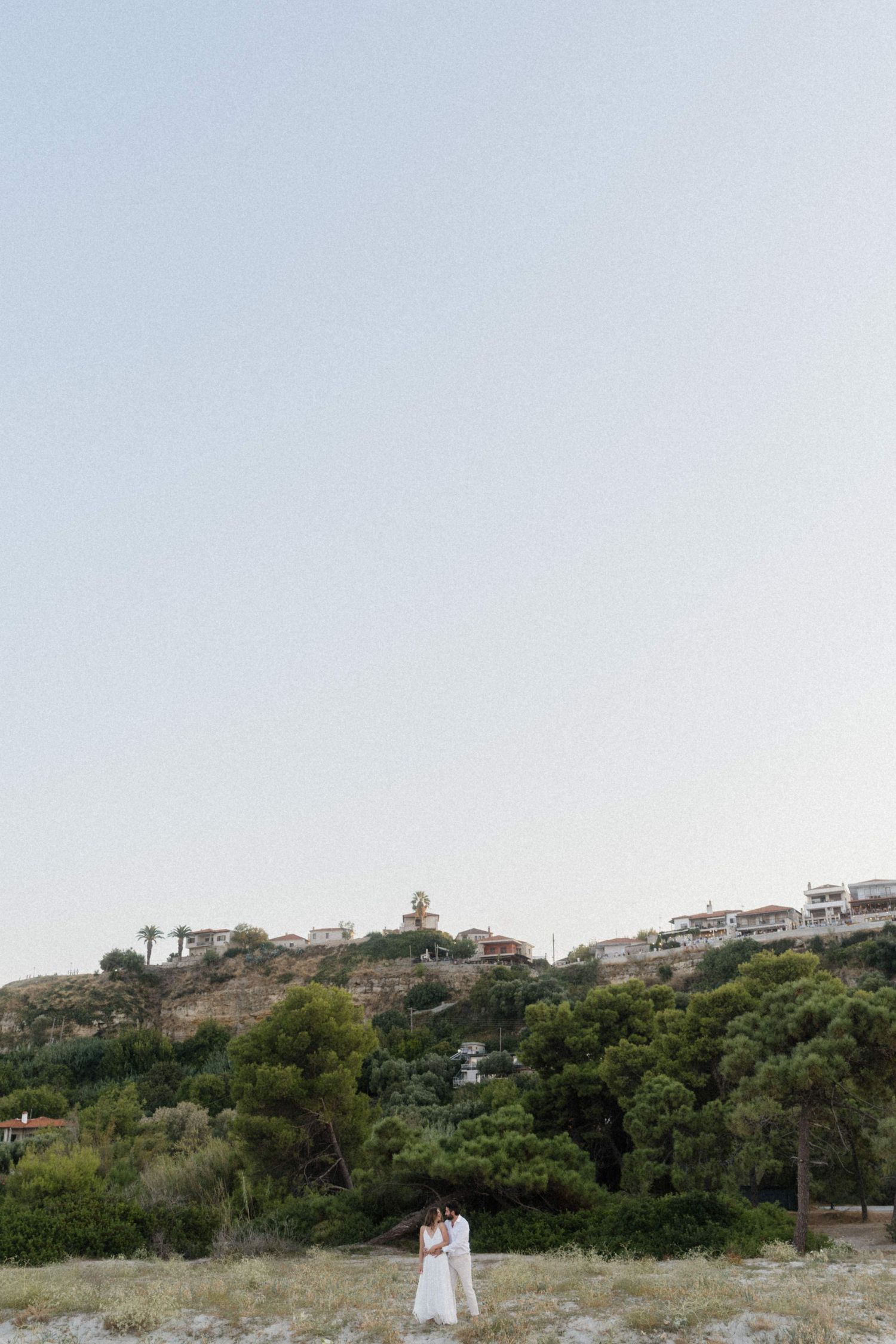 Couple in white embracing on a hillside with lush trees and Mediterranean village buildings under a clear sky.