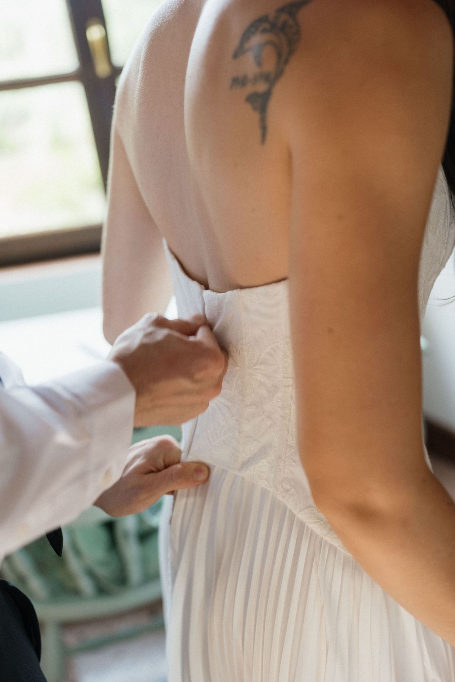 Bride with a shoulder tattoo in a strapless white pleated wedding gown as hands lace up the back of her dress.