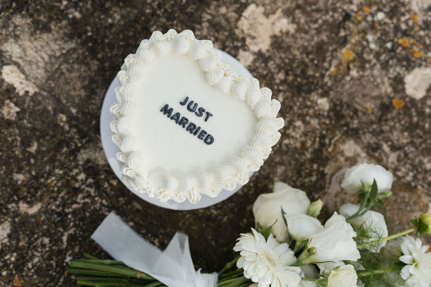 Heart-shaped white wedding cake topped with Just Married lettering, placed on a stone surface beside white roses and a ribbon.