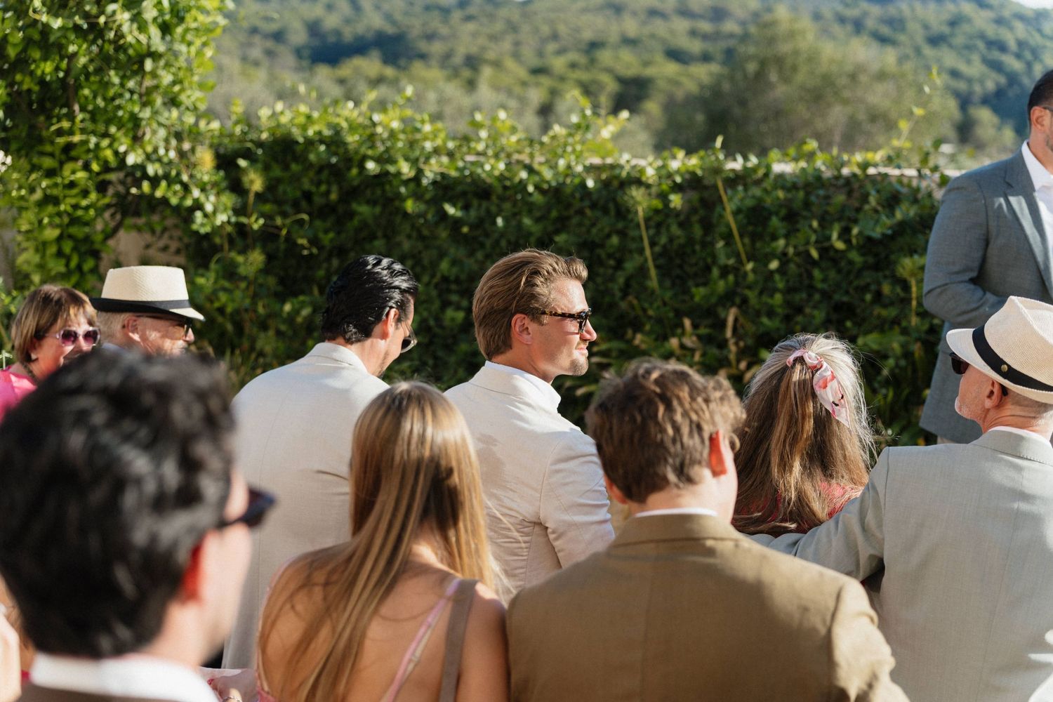 Wedding guests in summer attire sit facing an outdoor ceremony, with a green hedge and wooded hills in the background.