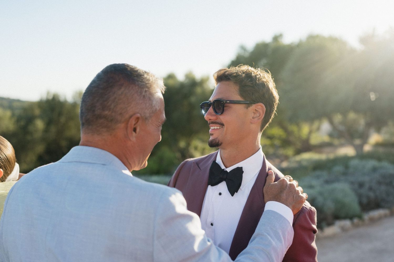 Smiling groom in sunglasses, burgundy jacket and bow tie shares a warm embrace with an older man at a sunny outdoor wedding.