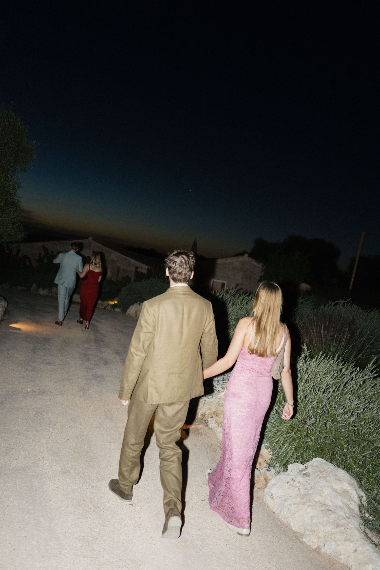 Wedding guests in pink and olive outfits walk along a gravel path at dusk under a dark blue evening sky.