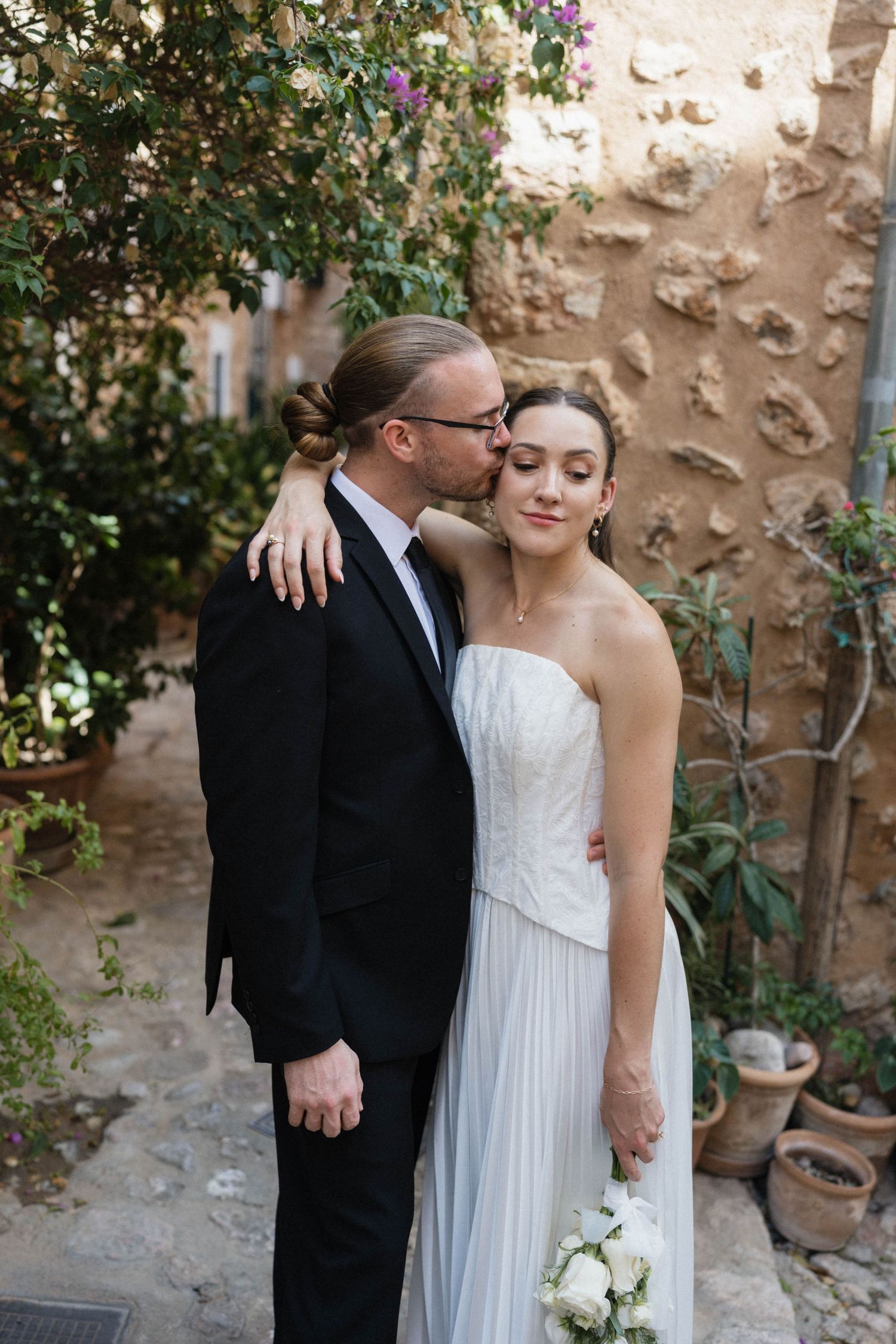 Groom in a black suit and glasses kisses his bride's forehead, she holds a white bouquet in a strapless gown by a rustic stone wall.