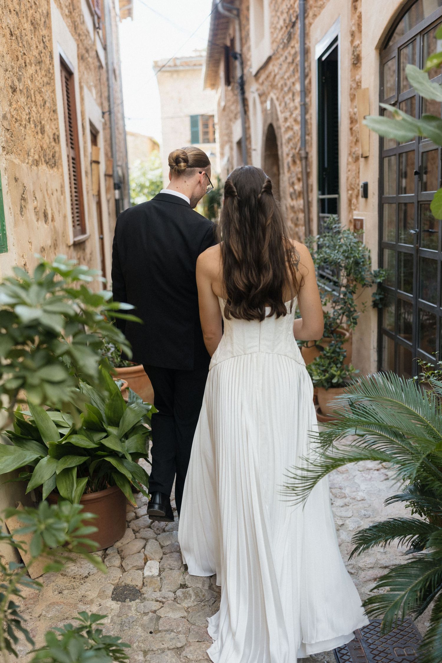 Bride in a pleated white gown walks with a groom in a dark suit along a cobblestone path past potted plants and stone buildings.