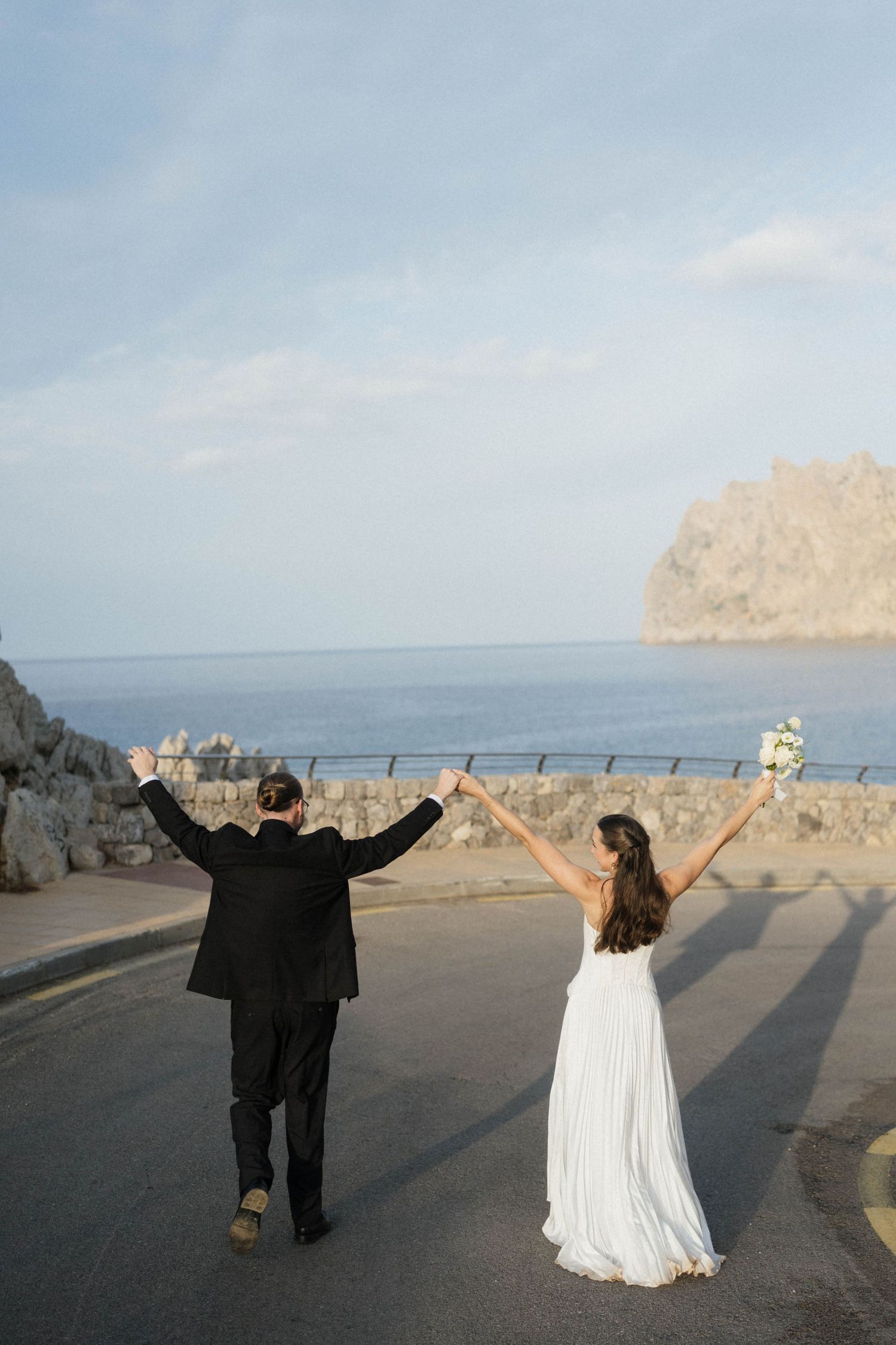 Newlyweds hold hands raised in celebration while walking along a seaside road with a bouquet and a rocky headland ahead.